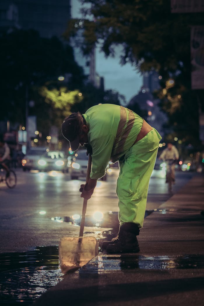 A city worker in bright attire cleans the street at dusk in an urban setting.
