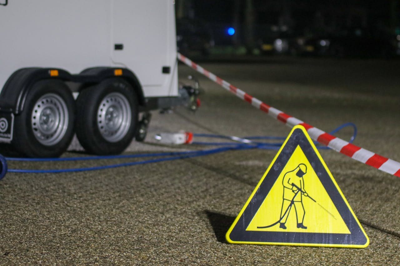 A construction warning sign and caution tape at night near a trailer, indicating ongoing work.