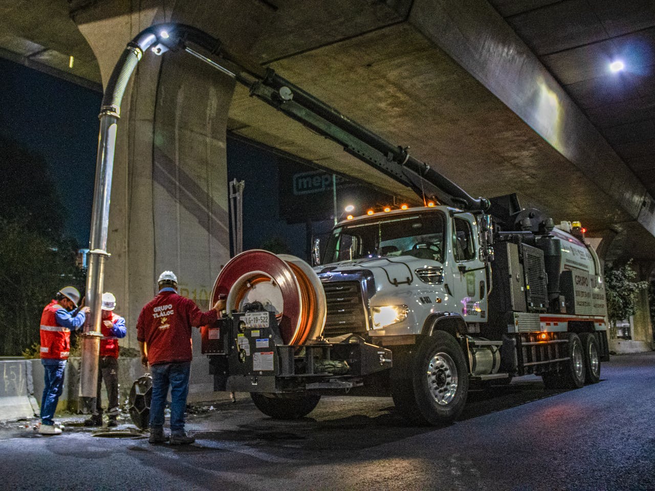 Workers operate a vacuum truck under a bridge at night for city maintenance.
